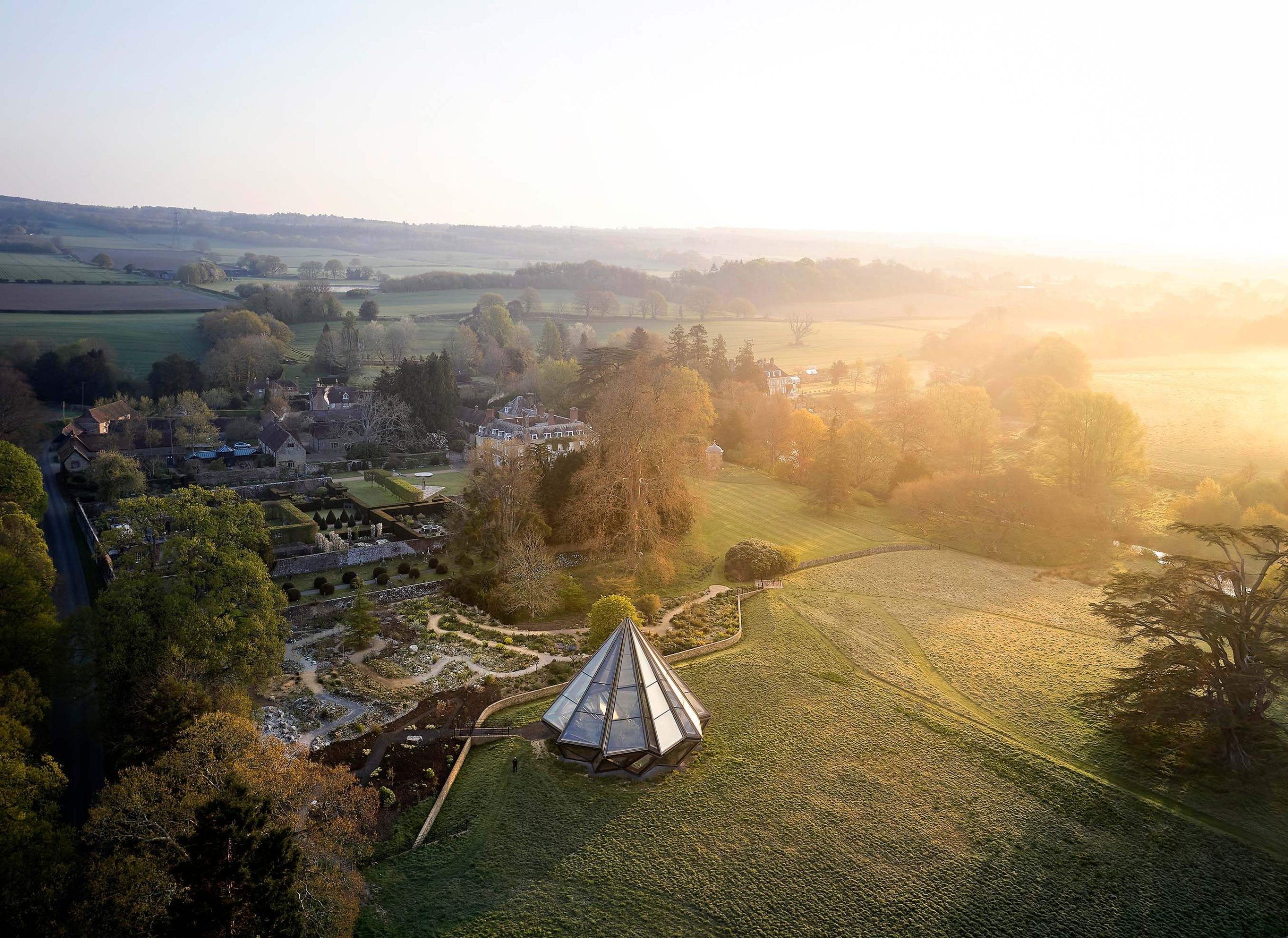 A modern glass greenhouse structure shaped like a diamond captured from above surrounded by gardens and fields