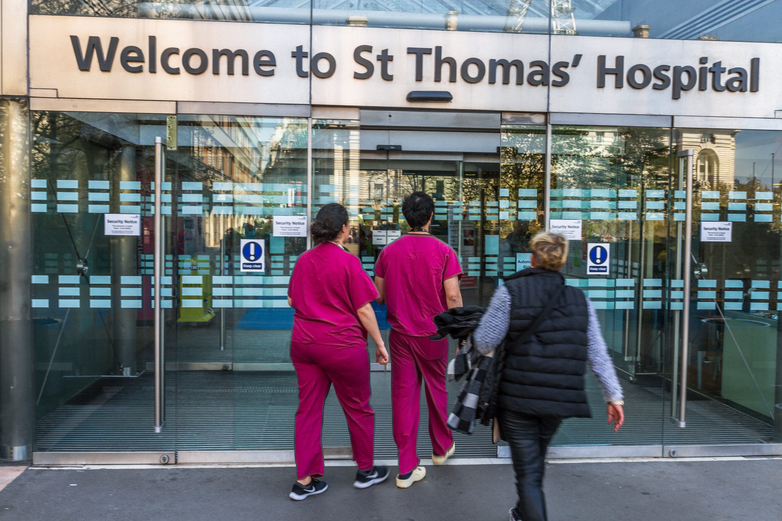 Glass door entrance with sign reading Welcome to St Thomas' Hospital. Two people in scrubs and one in casual clothing are entering the doorway
